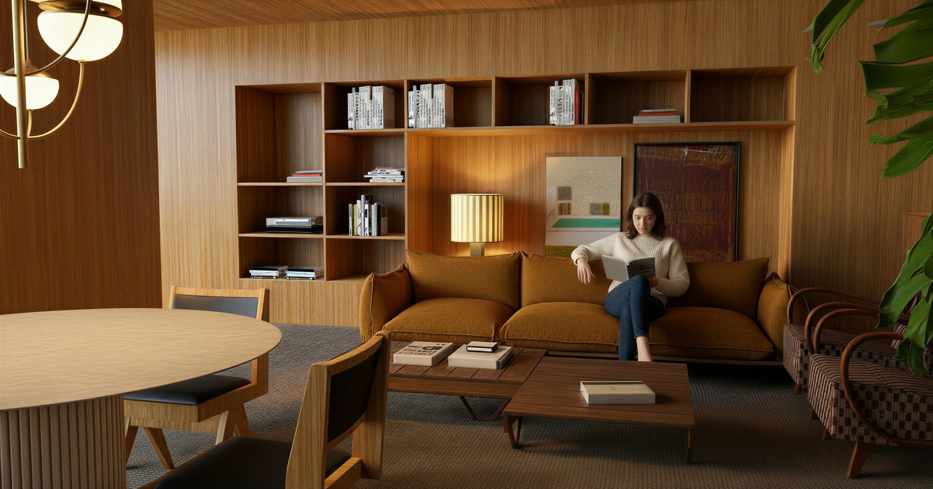 A woman sits on a brown sofa reading a book in a modern living room with wooden walls, a round table, and a bookshelf filled with books.