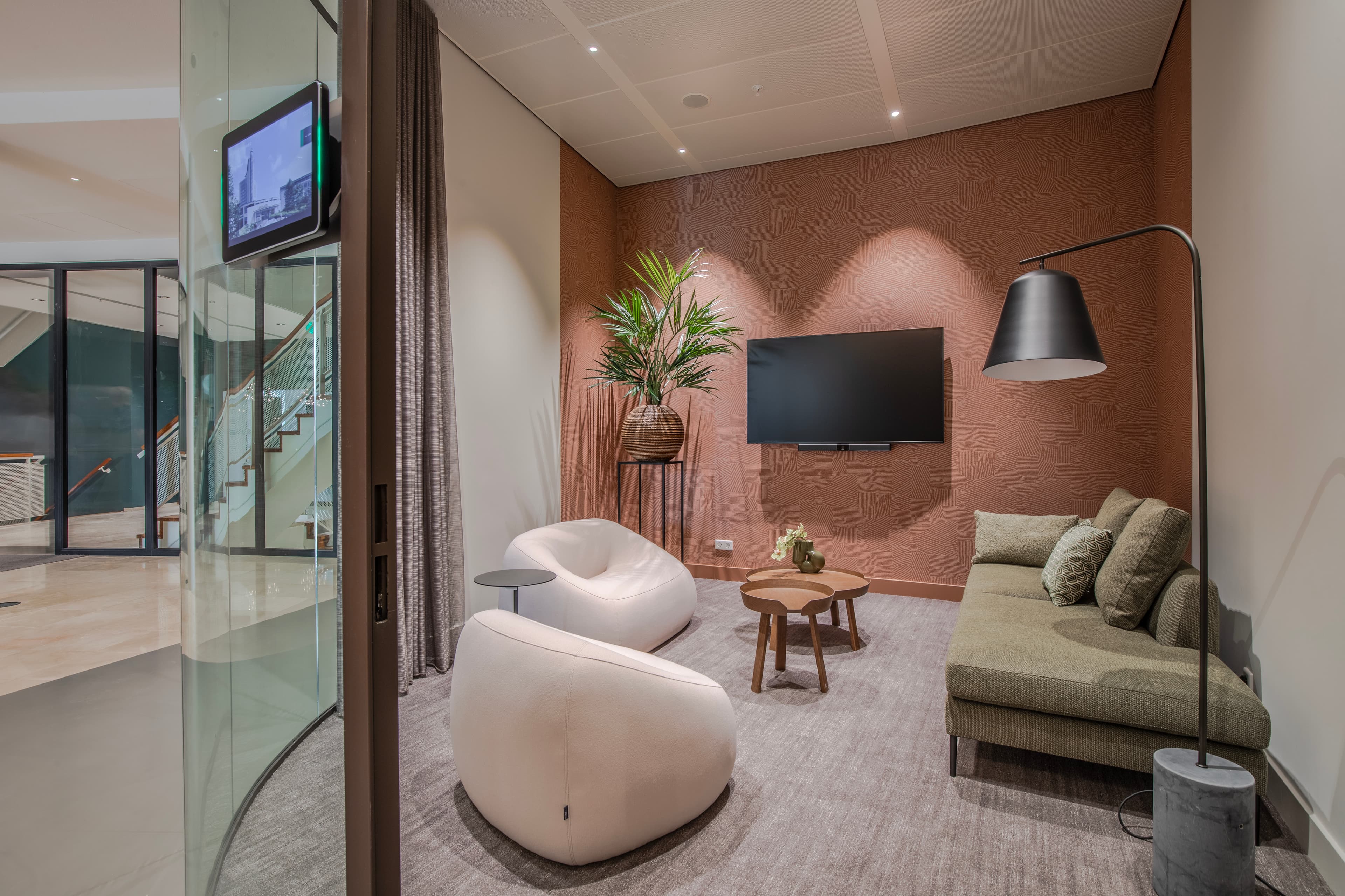 Modern lounge with beige bean chairs, a green sofa, flat-screen TV, tall potted plant, wooden coffee table, and floor lamp, all in a glass-enclosed room.