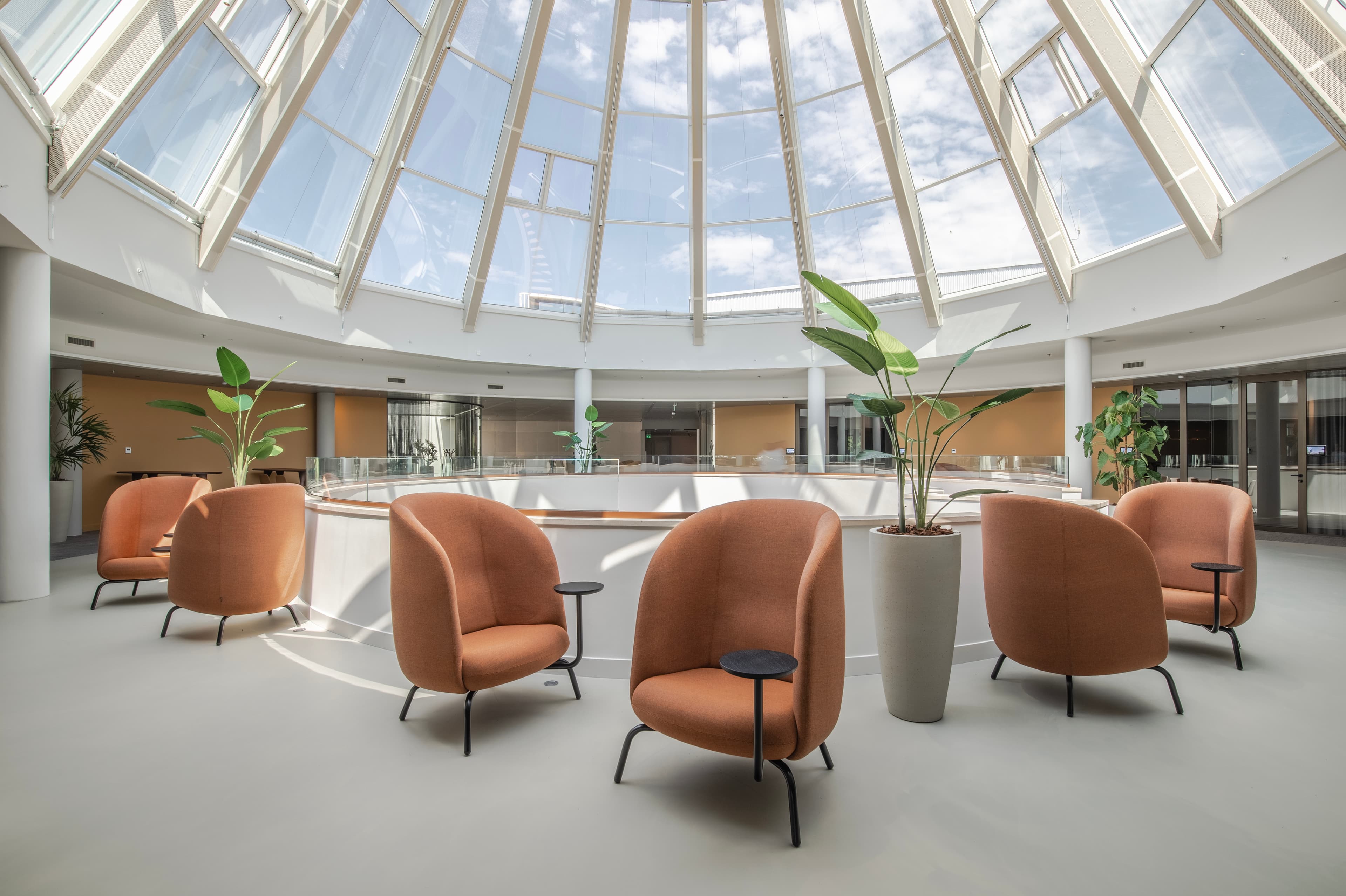 Spacious atrium with orange chairs and potted plants, beneath a large glass dome roof, creating a bright, modern, and inviting environment.