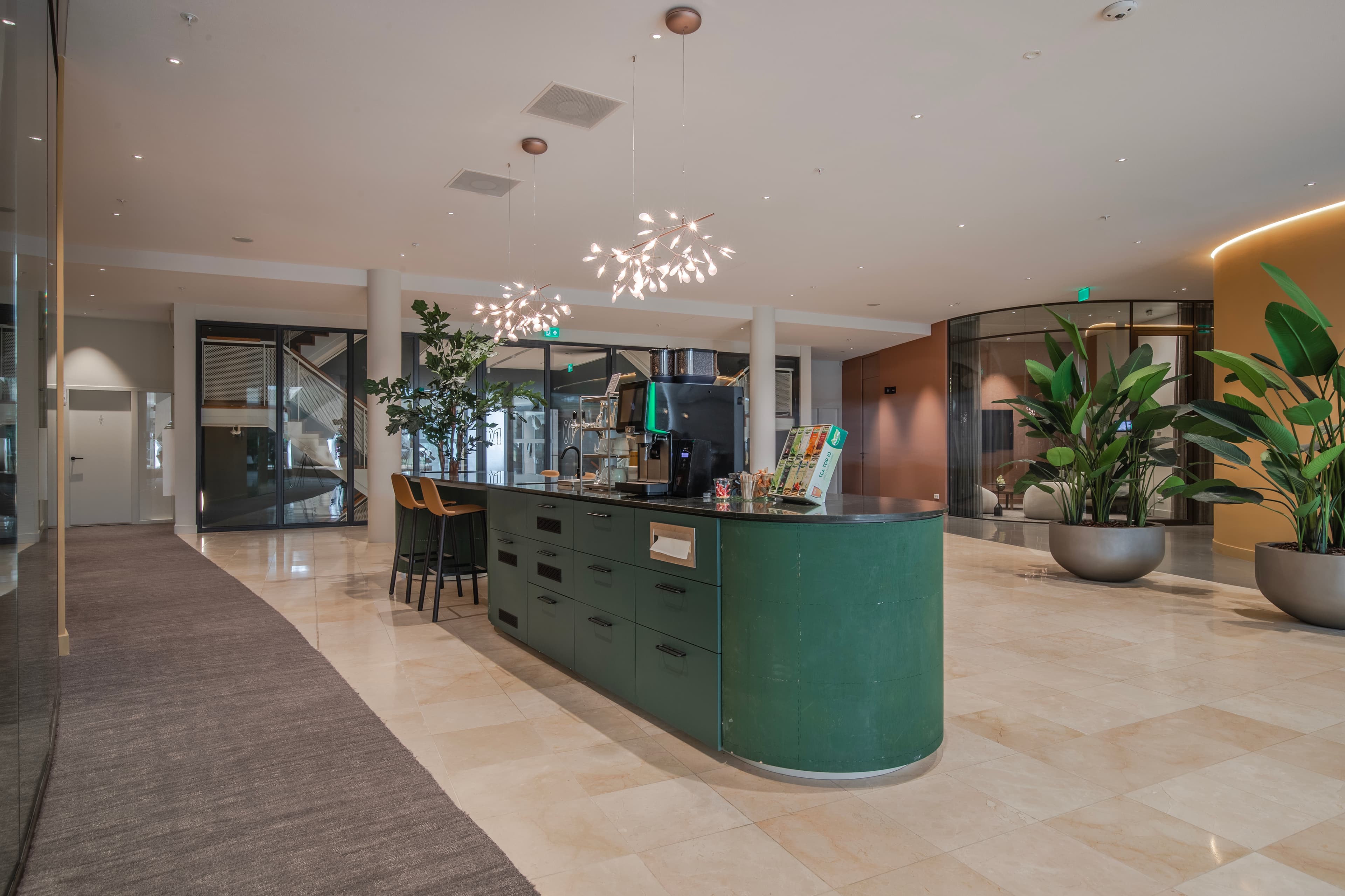 Modern lobby with a green reception desk, overhead pendant lights, potted plants, and beige tiled flooring leading to glass doors.