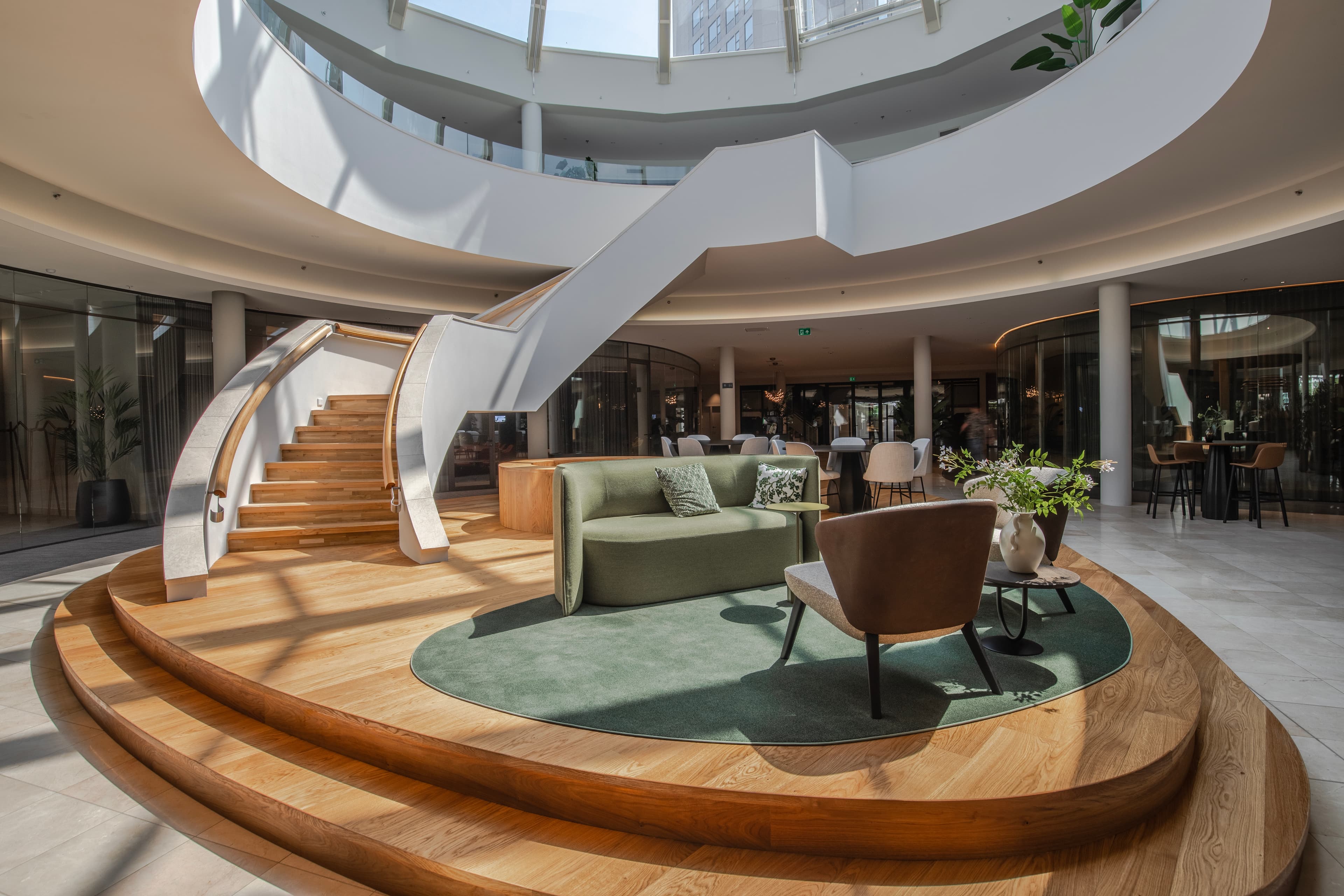 Spacious modern atrium with a curved staircase, wooden flooring, green couch, chairs, and plants under a skylight.