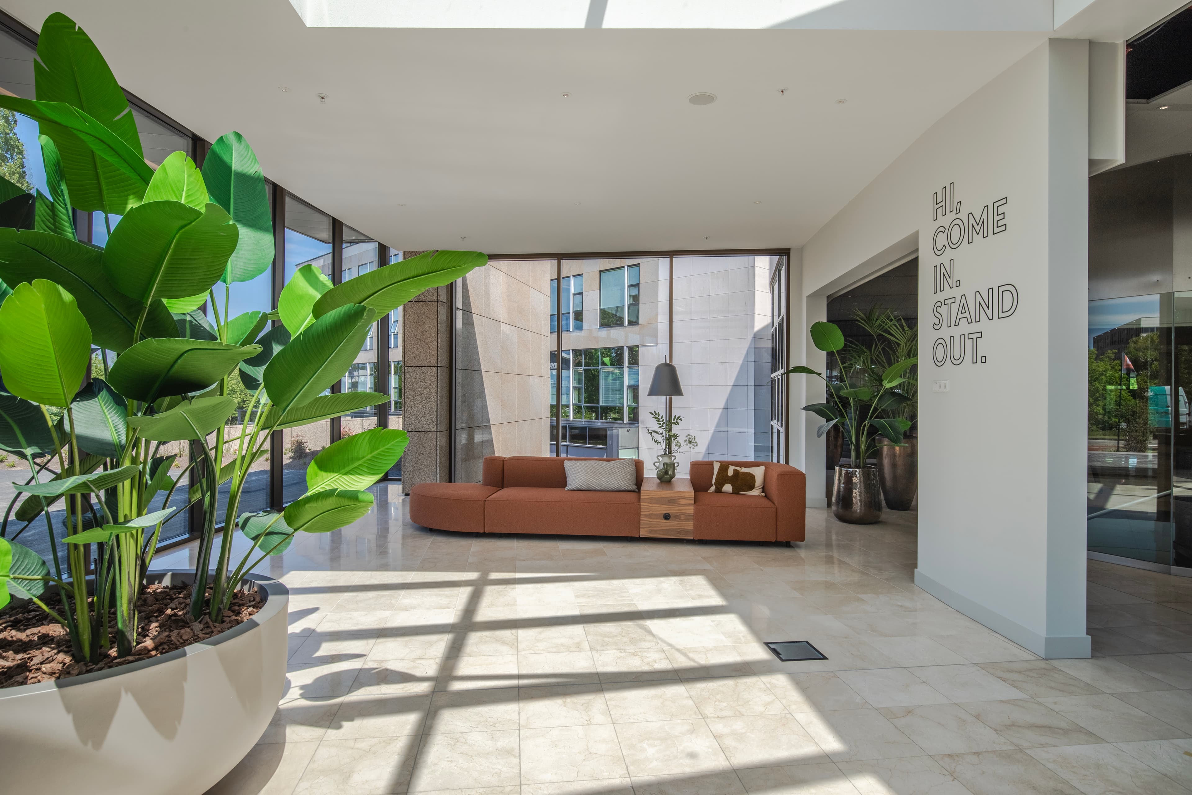 Modern lobby with large potted plants, sunlight streaming through glass walls, a brown sectional sofa, and a wall with "Hi. Come In. Stand Out." text.