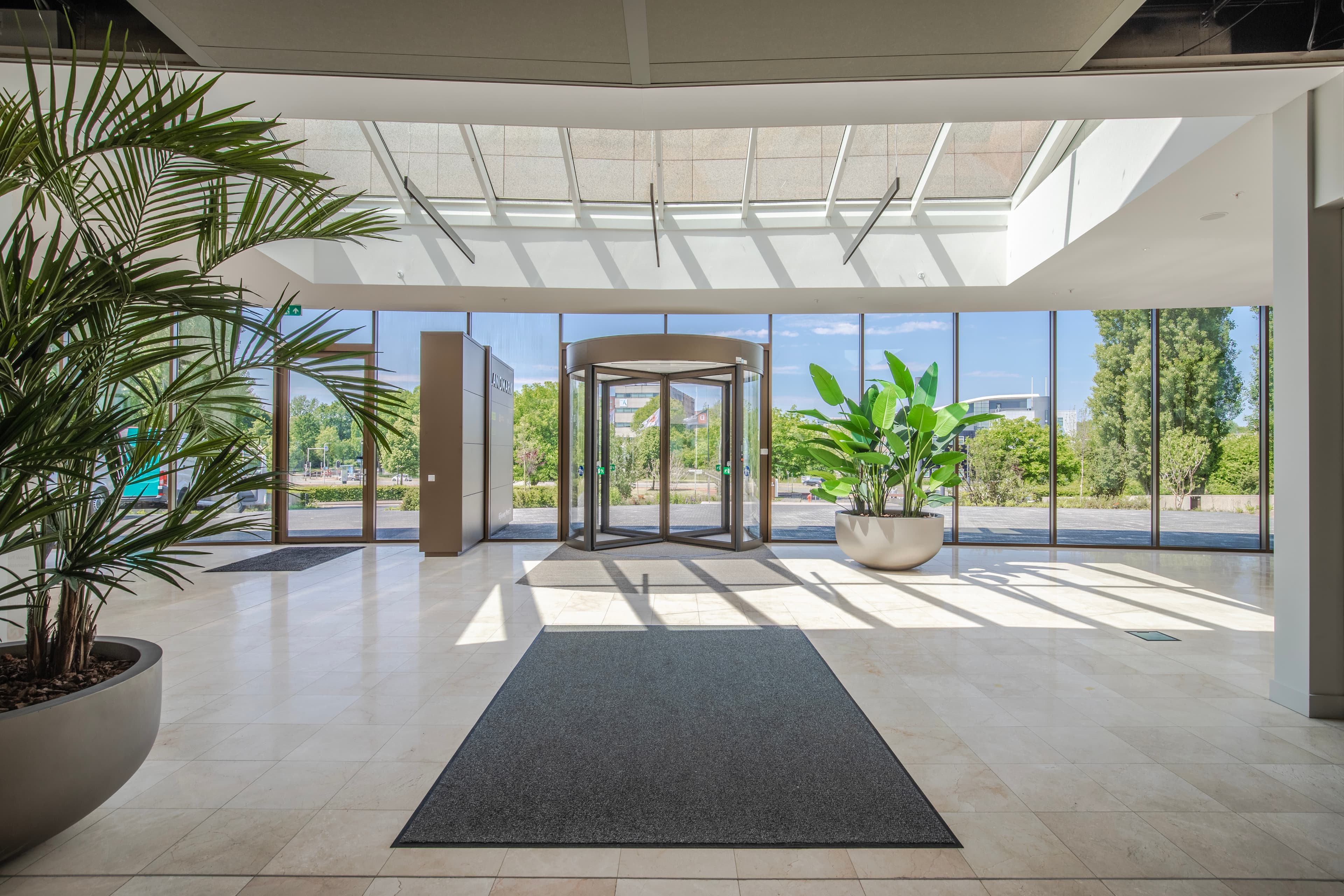 Spacious lobby with large windows, revolving door, tiled floor, floor mats, and potted plants, overlooking a green outdoor area.