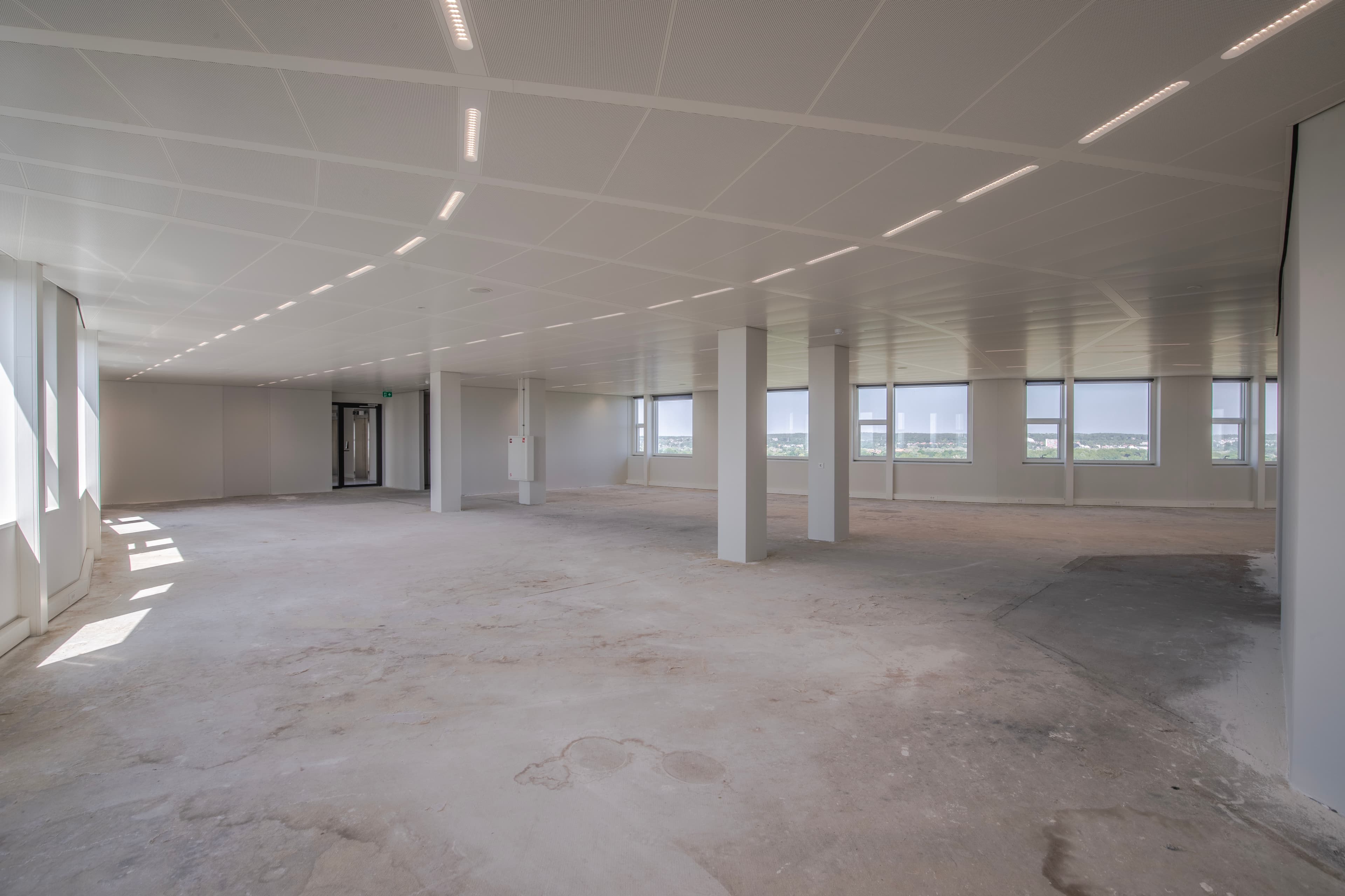 Spacious, empty room with unfinished flooring, large windows, and a modern ceiling with strip lighting. Natural light streams in through the windows.
