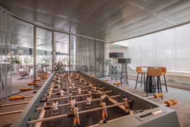 Modern office space with a foosball table in the foreground and a lounge area with plants and glass walls in the background.