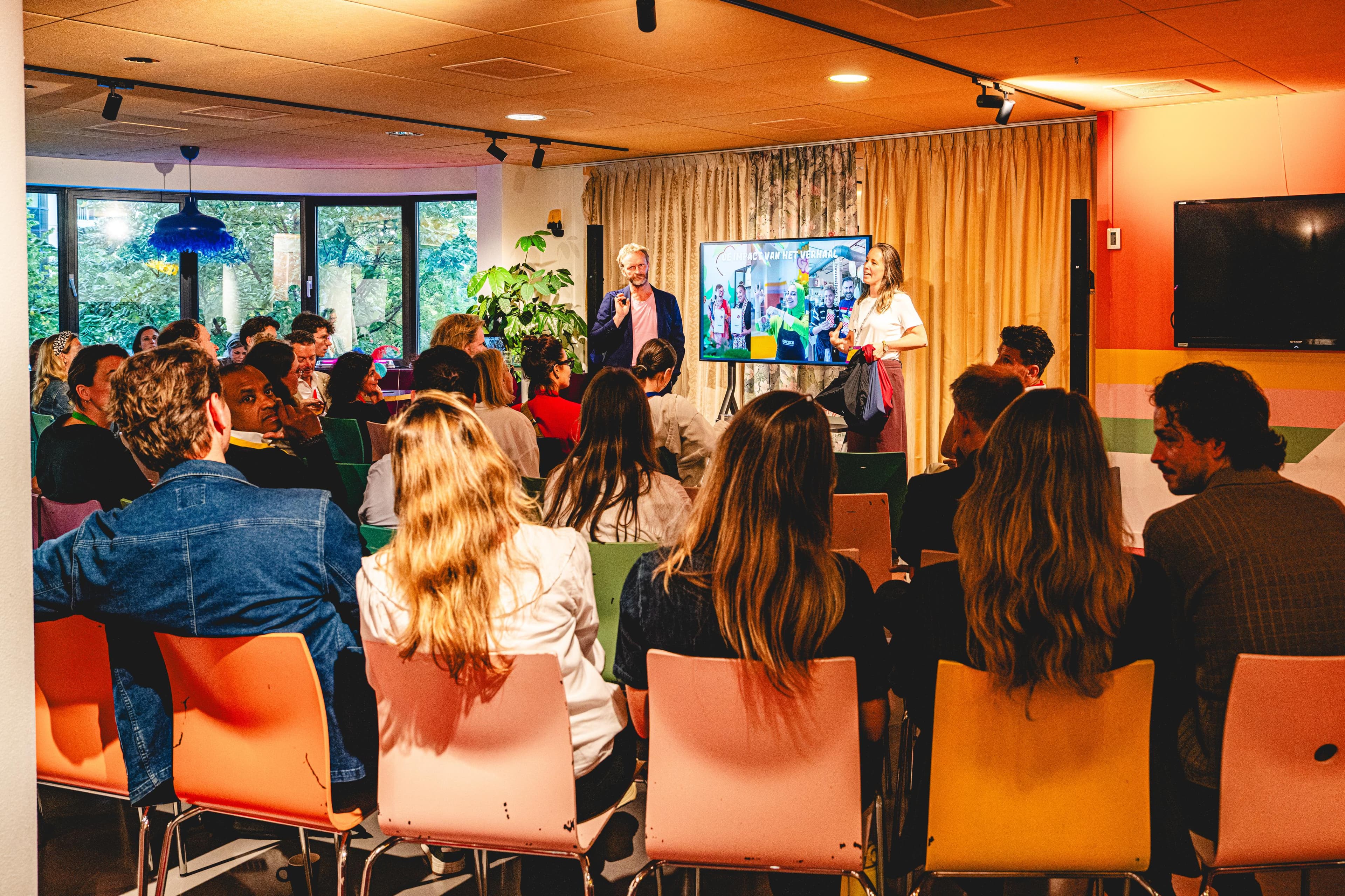 A speaker presents in front of an audience seated in colorful chairs, with a presentation screen in the background and large windows with trees outside.