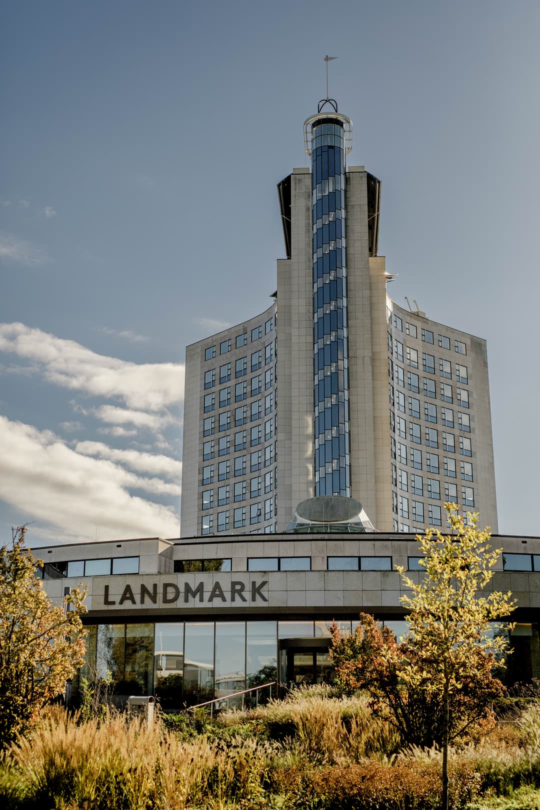 Modern building entrance with glass facade, revolving door, and people walking. Interior features stairs, plants, and framed pictures.