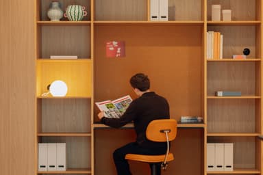 Person reading a magazine at a desk, surrounded by wooden shelves with books, vases, and a lit lamp.