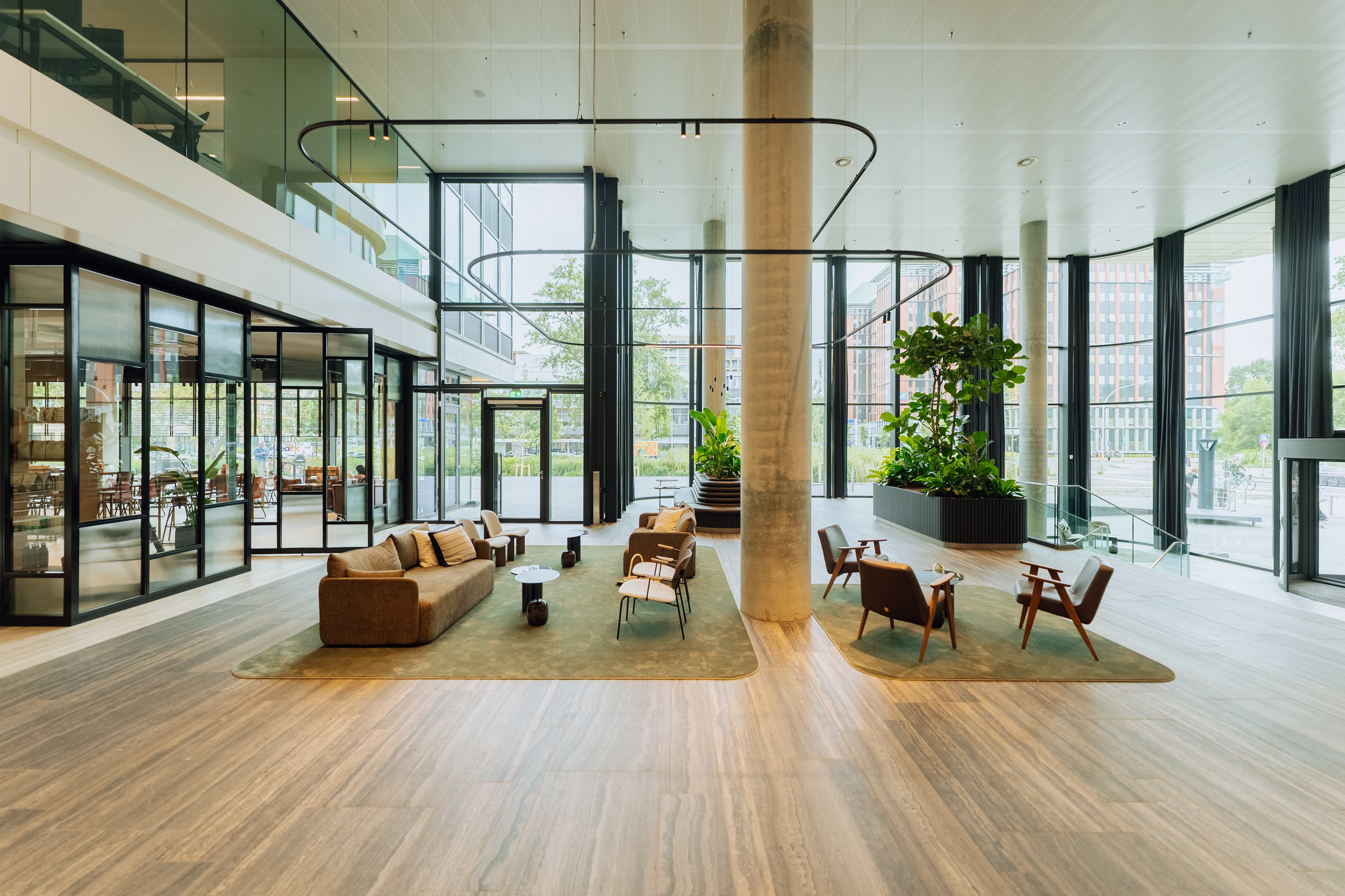 Modern lobby with brown sofas, chairs, and small tables on green rugs. Large windows, glass walls, and indoor plants enhance the open space.