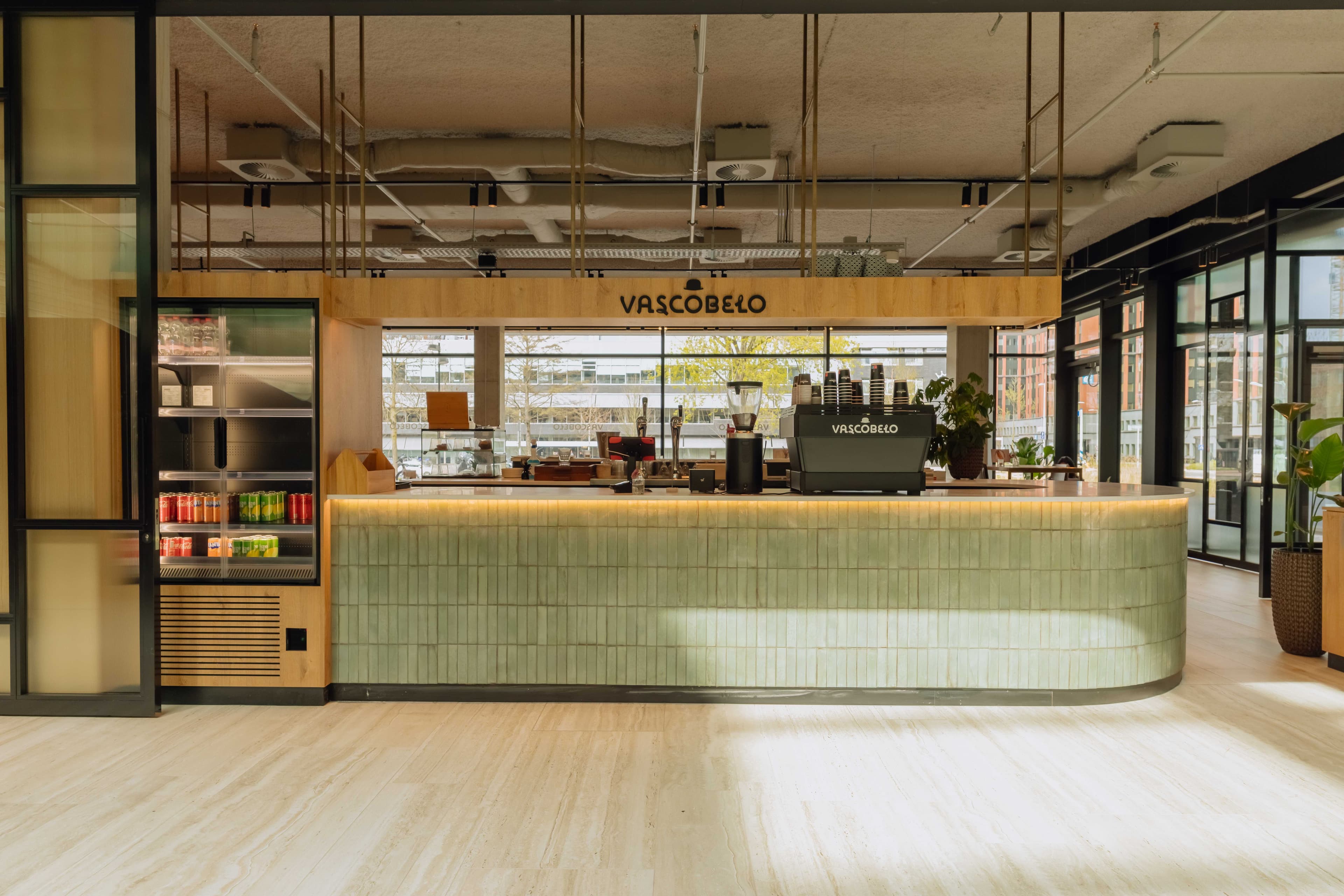 Modern coffee shop interior with a green-tiled counter, espresso machine, a fridge with drinks, and large windows in the background.