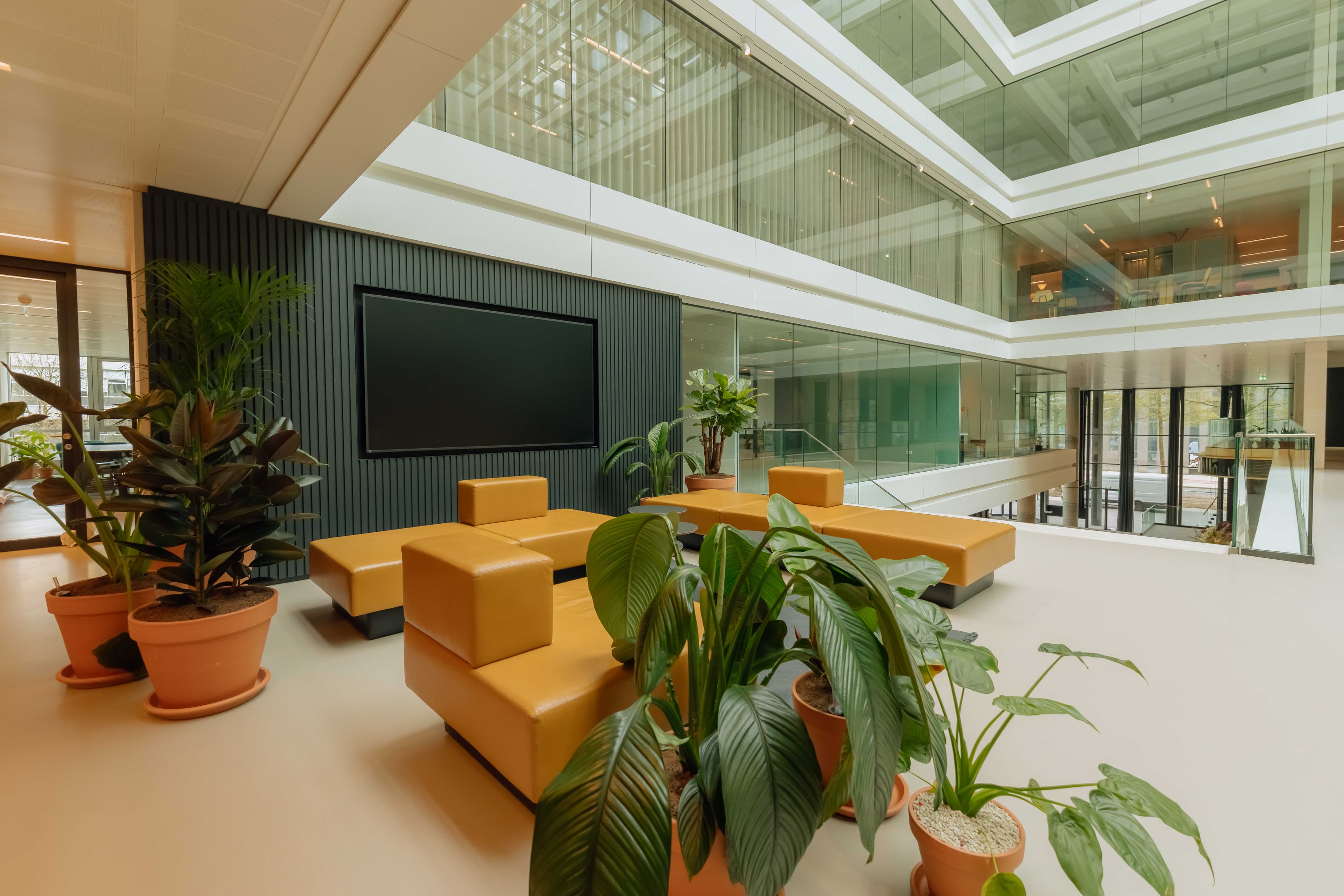 Modern atrium with orange seating, green potted plants, a large screen on a dark wall, and glass railings with offices visible above.