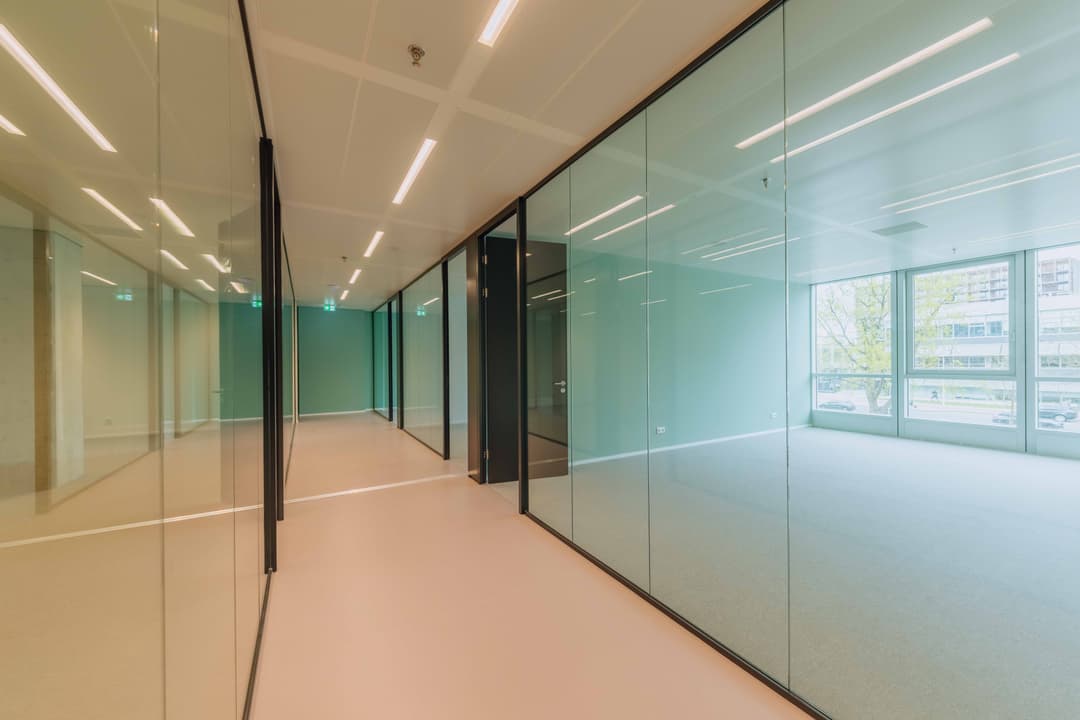 Modern office with glass walls, beige flooring, and ceiling lights. Large windows with city view and greenery outside.
