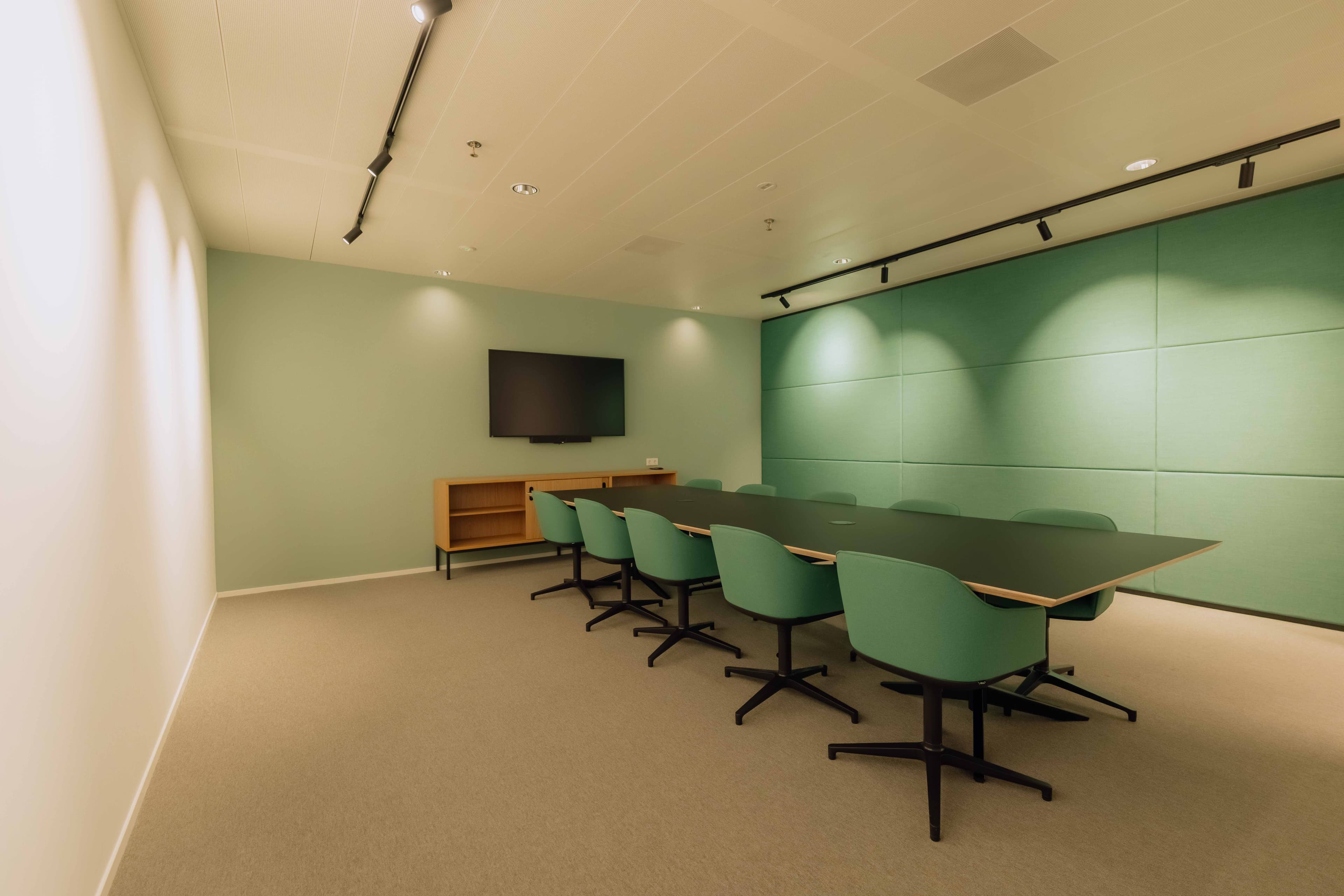 A modern conference room with a long table, eight green chairs, a wall-mounted TV, and minimalist decor in soft green tones.
