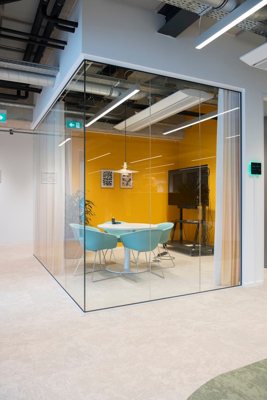 Modern meeting room with glass walls, yellow accent wall, round table, four blue chairs, pendant light, and plant in corner.