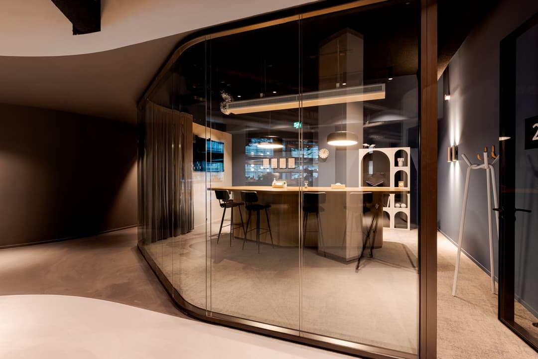 Modern office space with glass walls, a wooden table, high chairs, and ambient lighting. The interior has a sleek, contemporary design.