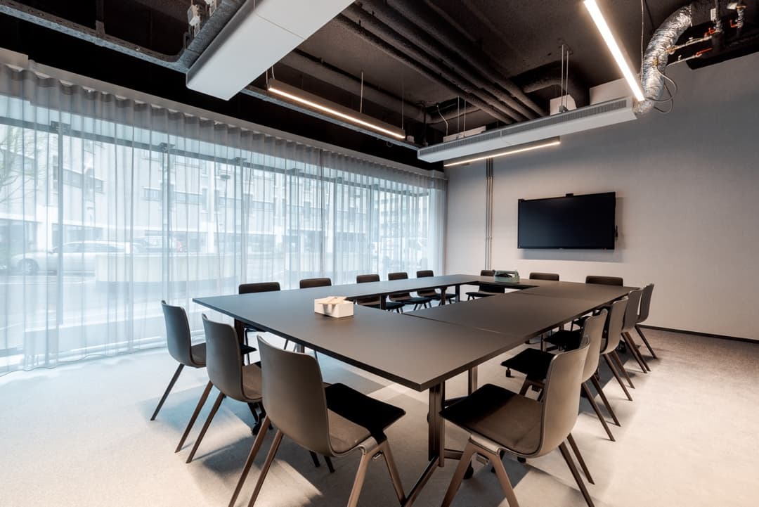 Modern conference room with a large black table, surrounded by chairs, a wall-mounted TV, and floor-to-ceiling windows with sheer curtains.