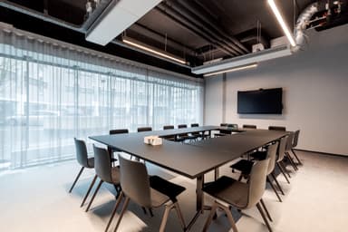 Modern conference room with a large black table, surrounded by chairs, a wall-mounted TV, and floor-to-ceiling windows with sheer curtains.