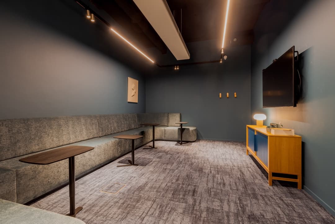 Modern lounge with blue walls, built-in seating, small tables, a wall-mounted TV, and a wooden console with a lamp.