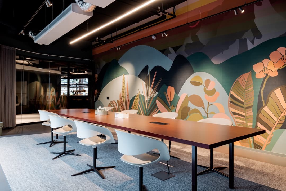 Modern meeting room with a long wooden table, white chairs, vibrant nature-themed mural, and a wall-mounted TV on a gray carpet.