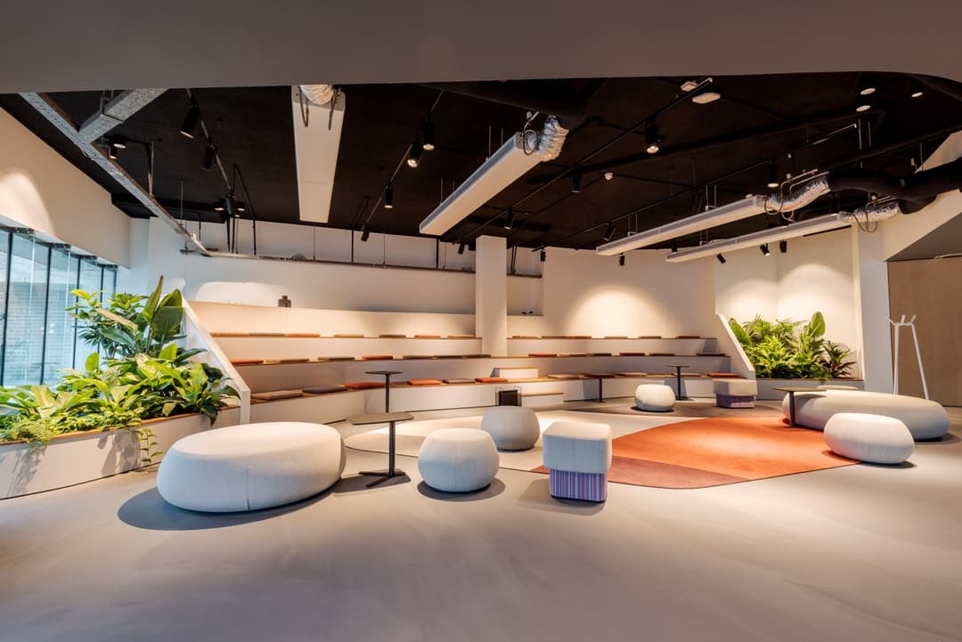 Modern interior with tiered seating, round cushioned stools, tables, and indoor plants. Large windows allow natural light to illuminate the space.