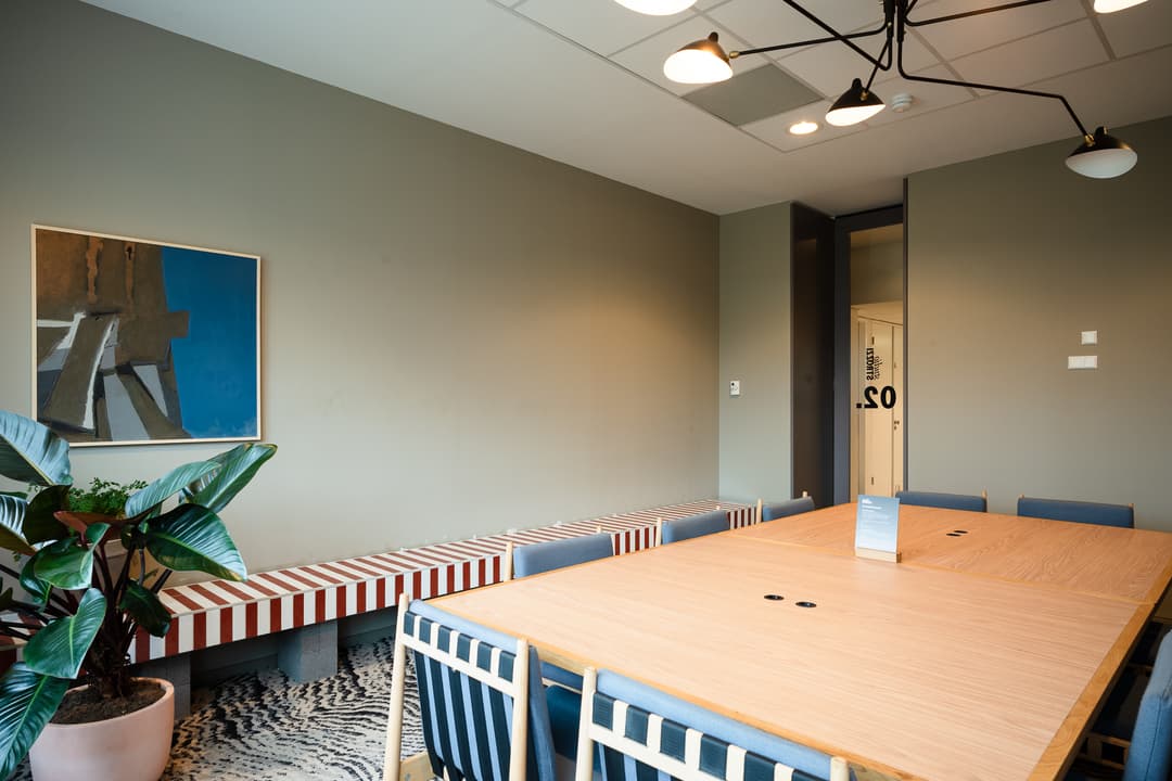 Modern meeting room with a wooden table, blue chairs, a striped bench, a painting on the wall, and a potted plant.