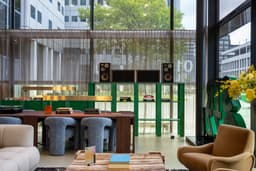 Modern lounge with plush seating, a wooden table, books, and speakers. Large windows reveal a view of urban greenery outside.