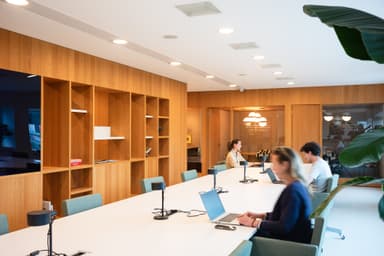 Modern office with wooden shelves, long white table, people working on laptops; soft lighting and a plant in the foreground.