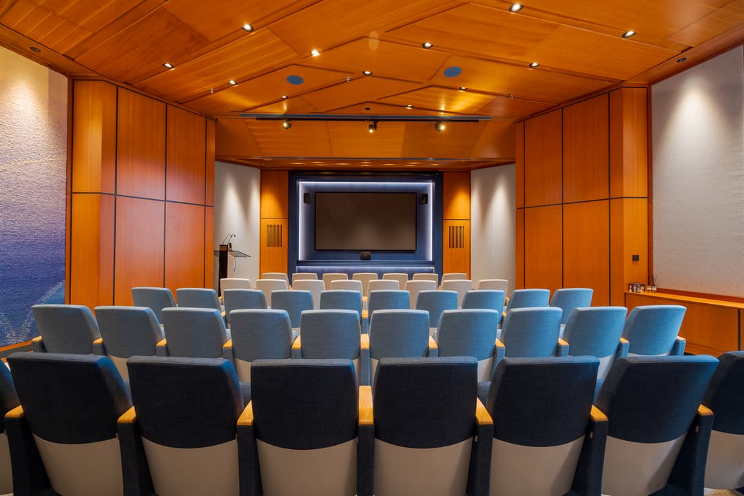 A small auditorium with blue and gray seats facing a large screen, surrounded by wooden paneling and soft lighting.