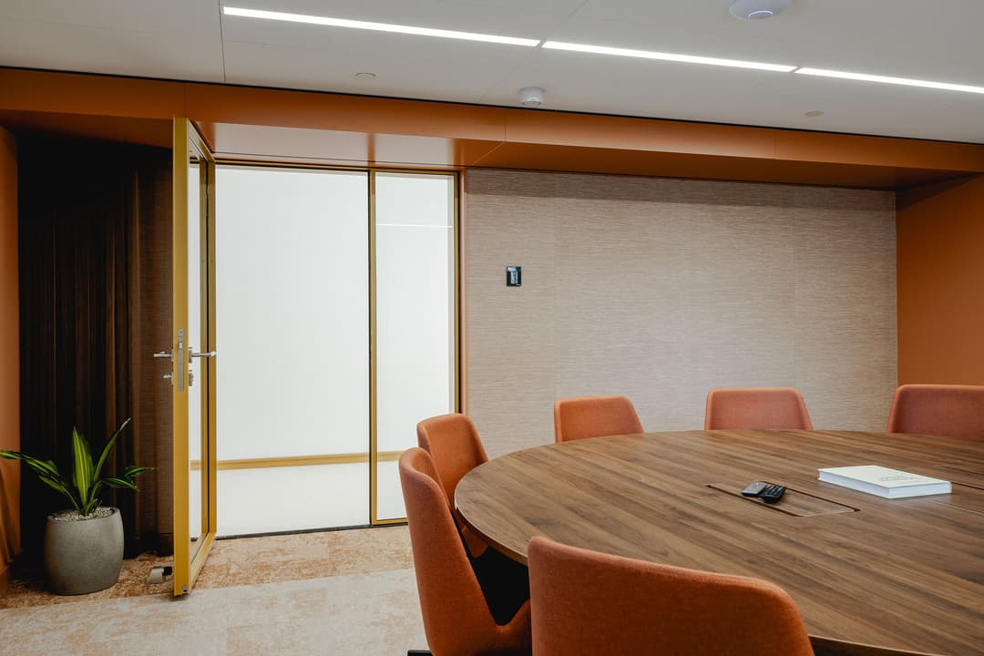 Modern conference room with a large wooden round table, orange chairs, an open door, and a potted plant in the corner.