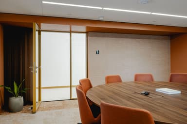 Modern conference room with a large wooden round table, orange chairs, an open door, and a potted plant in the corner.