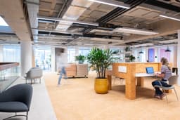 Modern open-plan office with plants, a person sitting at a desk with a laptop, and another person walking by. Bright and spacious environment.