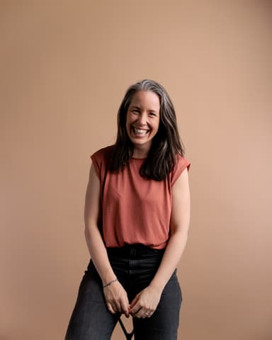 A person with long hair smiles broadly, wearing a sleeveless rust-colored top and dark pants, set against a plain beige background.