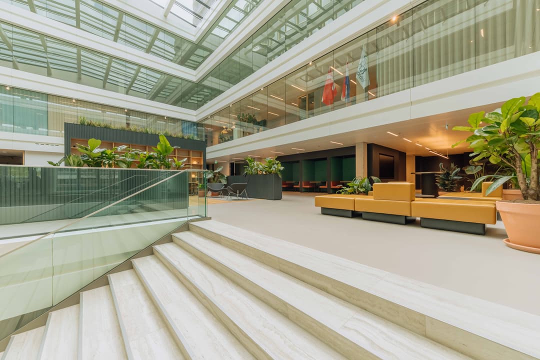 Modern atrium with glass walls, indoor plants, orange seating, and a staircase; flags displayed inside the spacious, well-lit area.