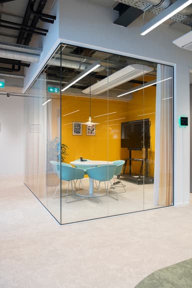 Modern meeting room with glass walls, yellow accent wall, round table, four blue chairs, pendant light, and plant in corner.